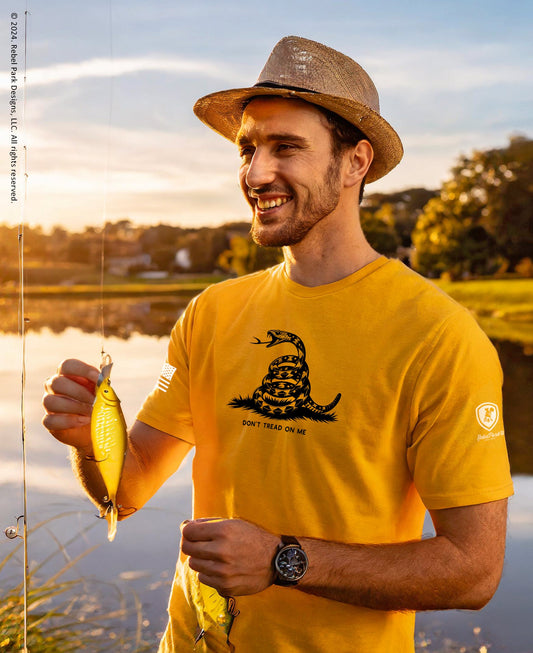 Man in yellow shirt with snake design holding a fishing lure by a lake.