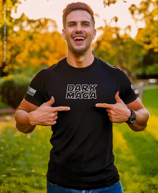 Man wearing a black t-shirt with 'DARK MAGA' text, pointing at it outdoors.