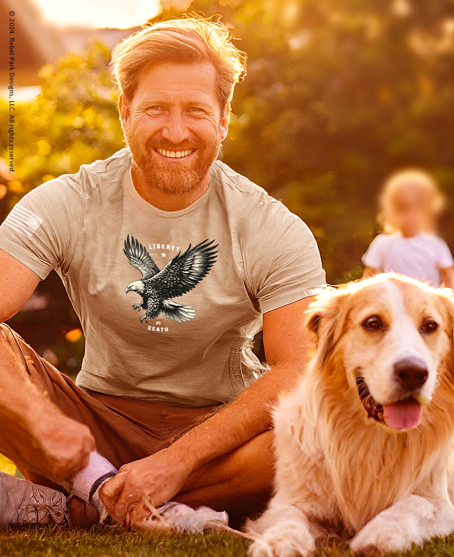 Man sitting on grass with a dog, wearing a t-shirt with an eagle design.