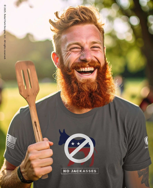 Man with a beard holding a wooden spatula, wearing a t-shirt with a logo and text, outdoors.