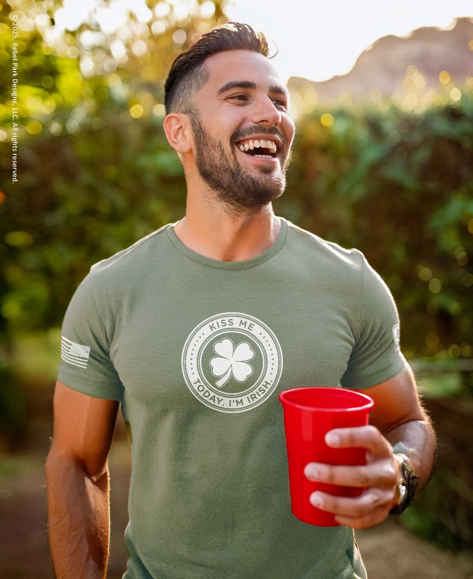 Man wearing a green t-shirt with a shamrock design and text, holding a red cup outdoors.