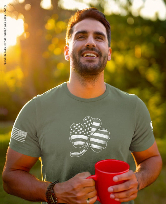 Man wearing a green t-shirt with a shamrock design, holding a red mug outdoors.