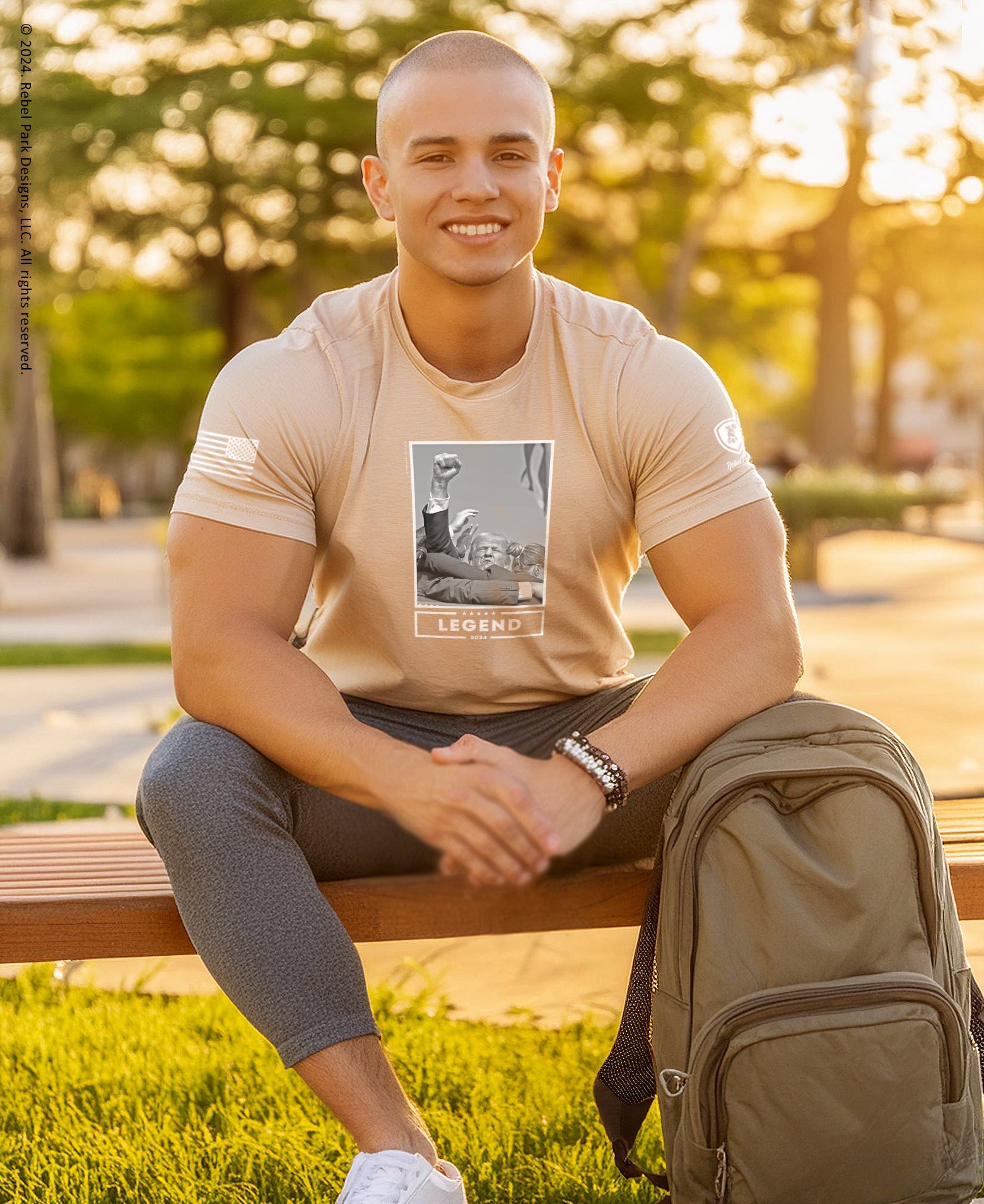 Man sitting on a bench in a park with a backpack, wearing a beige t-shirt with a graphic design.