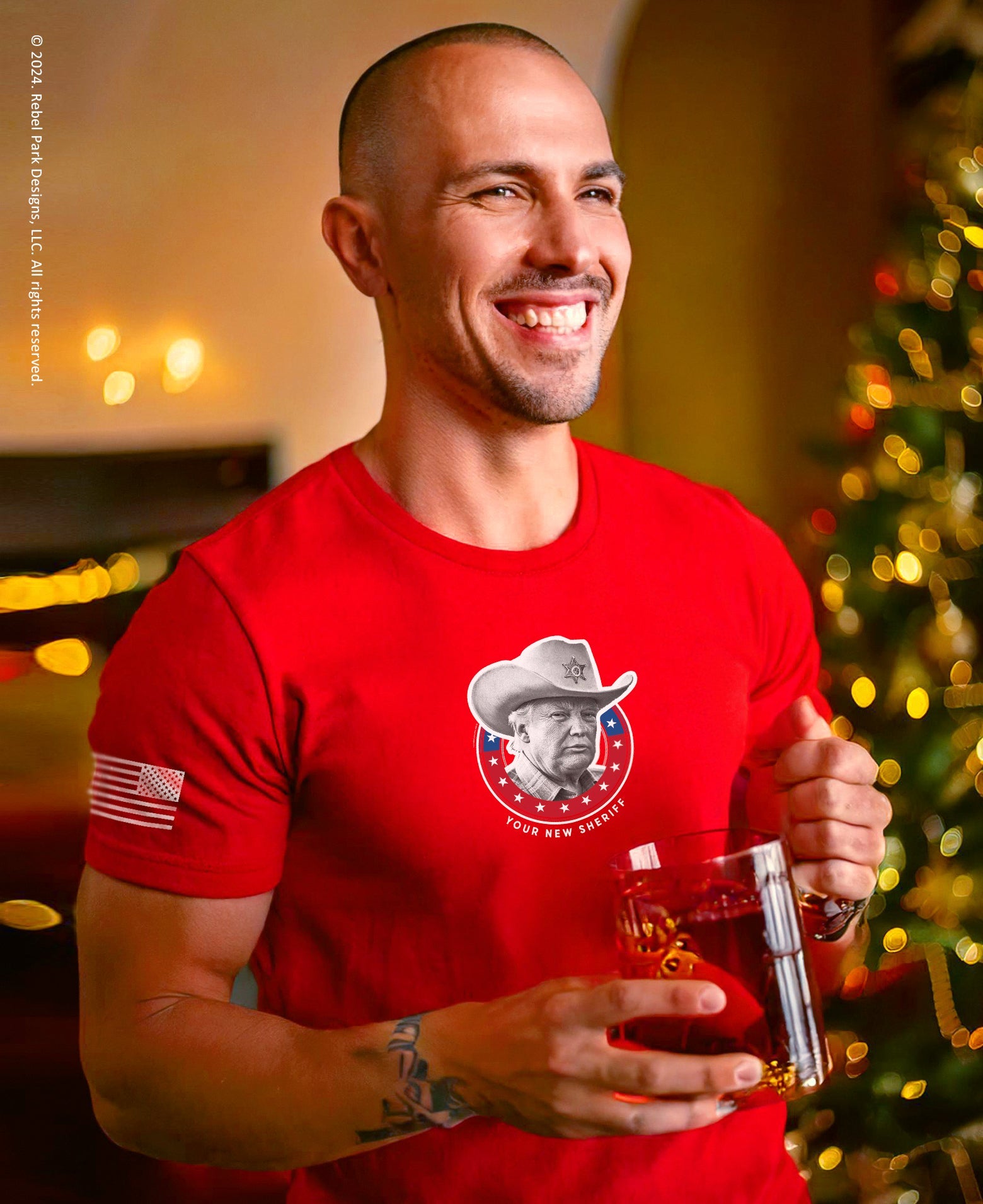 Man wearing a red t-shirt with a logo, holding a glass of wine, in a festive indoor setting.