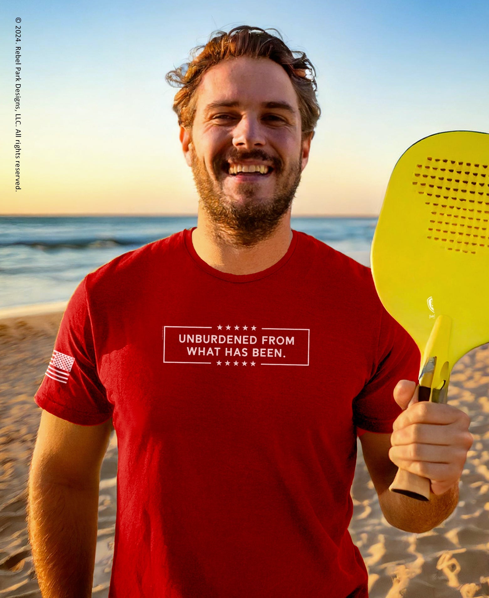 Man holding a yellow paddle on a beach with a red t-shirt displaying text.