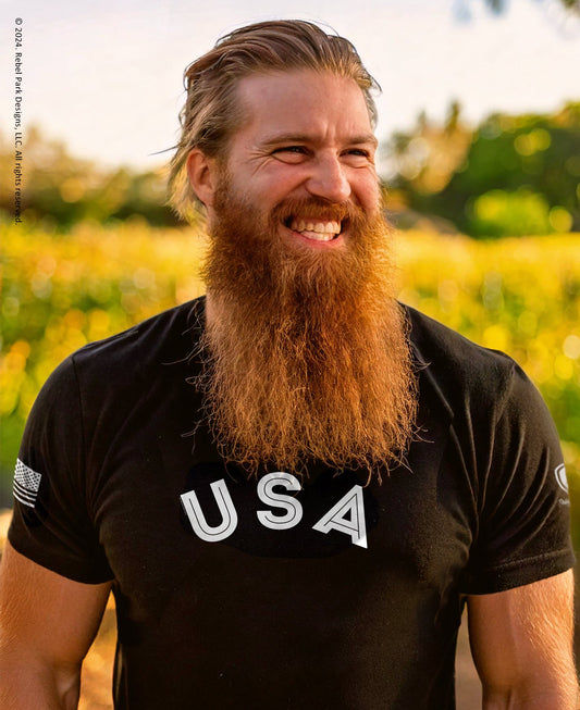 Man with a long beard wearing a black 'USA' t-shirt in a field.