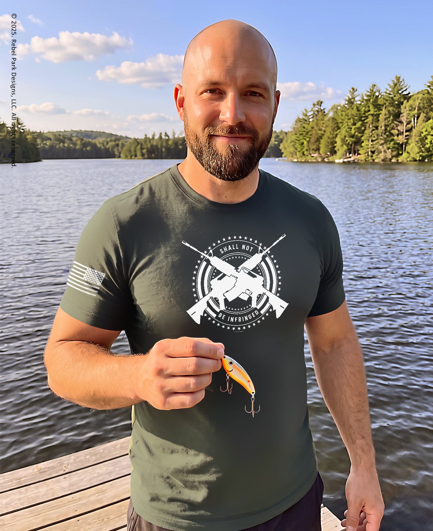 A man standing on a wooden dock by a lake wearing an olive green t-shirt with a white graphic print that reads 'WILD LIFE LURE COMPANY'. He is holding a yellow and orange fishing lure.