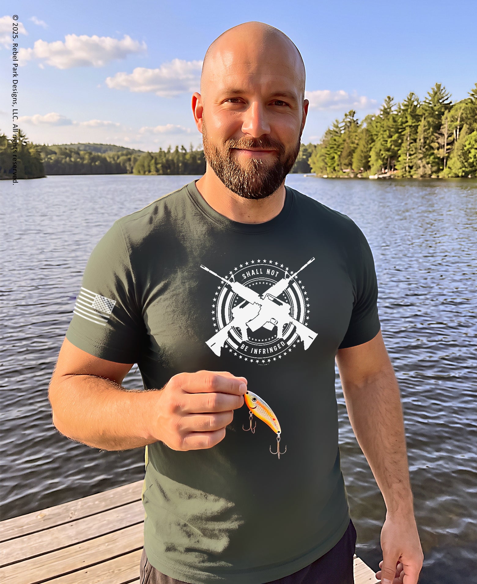 A man standing on a wooden dock by a lake wearing an olive green t-shirt with a white graphic print that reads 'WILD LIFE LURE COMPANY'. He is holding a yellow and orange fishing lure.