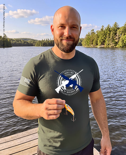 A man standing on a wooden dock by a lake wearing an olive green t-shirt with a blue and white military-themed graphic. He is holding a yellow and orange fishing lure.