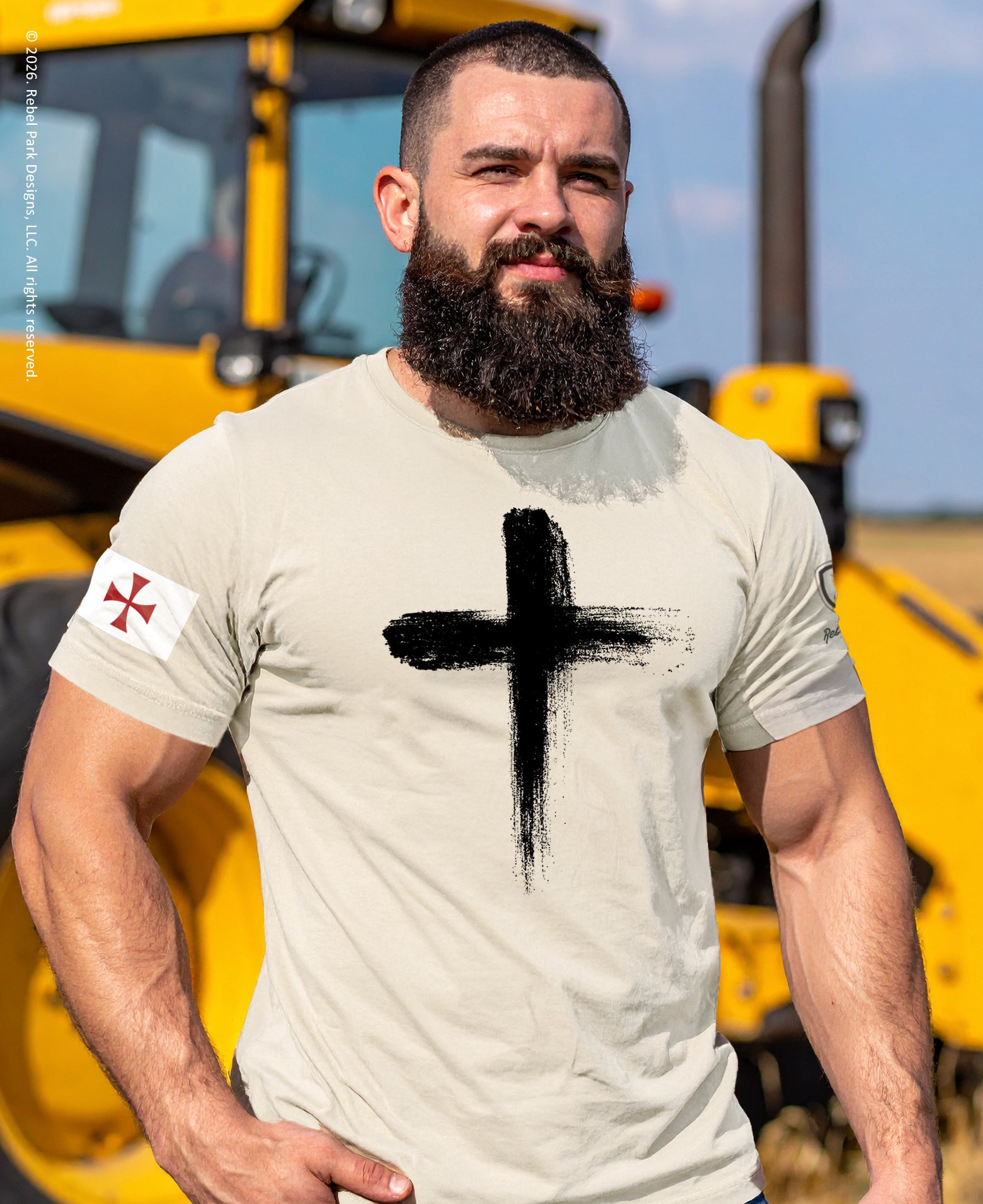 Man wearing a t-shirt with a black cross design in front of a yellow bulldozer.