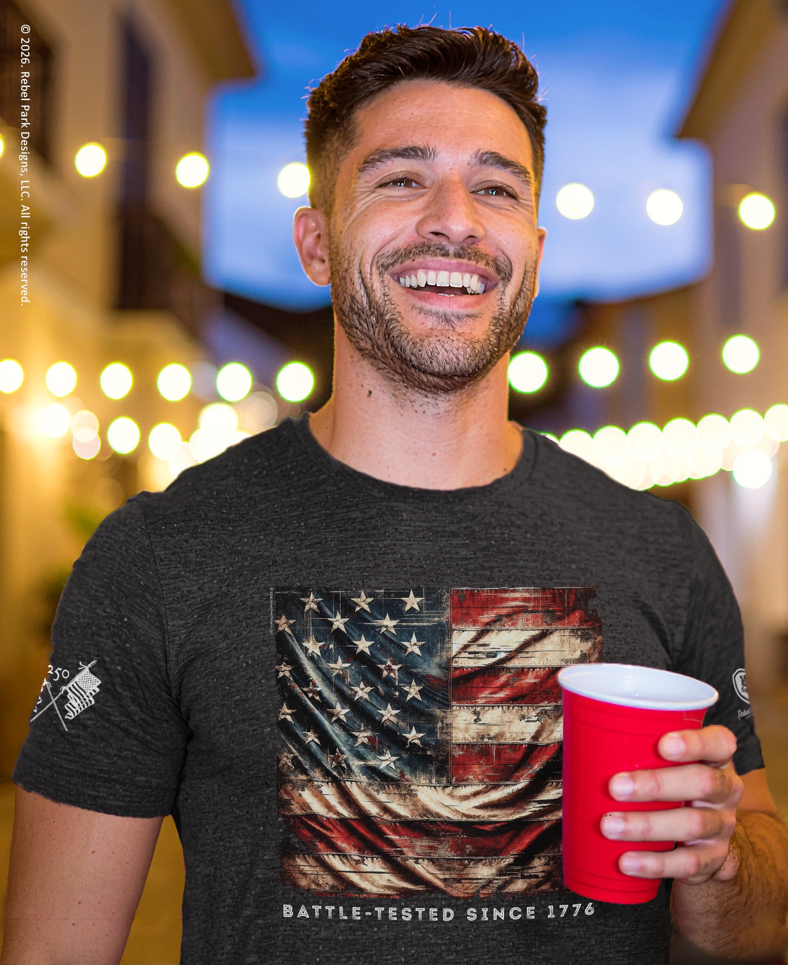Man wearing a t-shirt with an American flag design, holding a red cup, against a blurred background with lights.