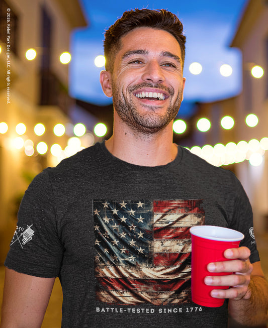 Man wearing a t-shirt with an American flag design, holding a red cup, against a blurred background with lights.