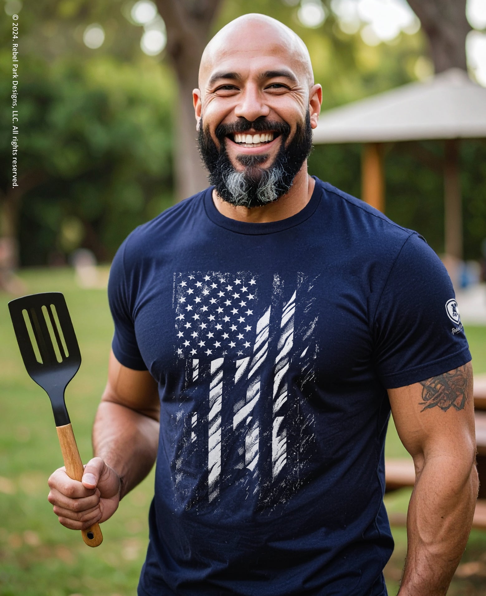 Man wearing a navy blue t-shirt with an American flag design, holding a spatula outdoors.