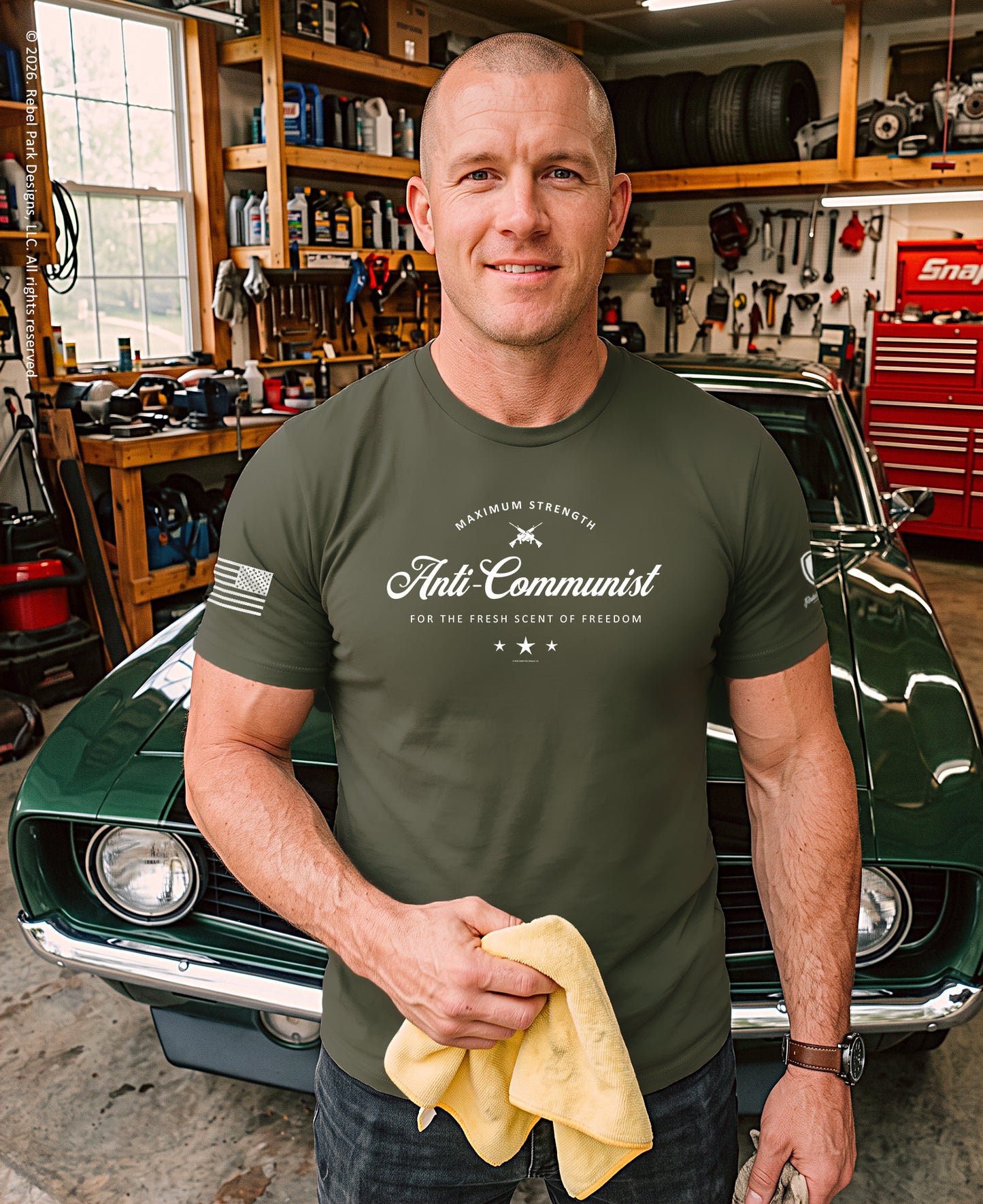 A man in a garage stands in front of a green classic car, wearing an olive green t-shirt with a white graphic that reads 'Anti-Communist' and holding a yellow microfiber cloth.