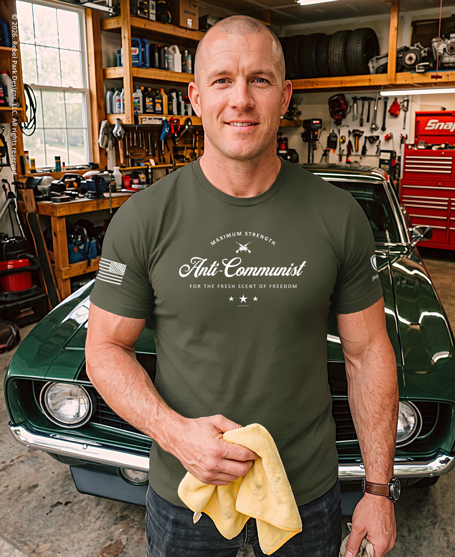 A man in a garage stands in front of a green classic car, wearing an olive green t-shirt with a white graphic that reads 'Anti-Communist' and holding a yellow microfiber cloth.