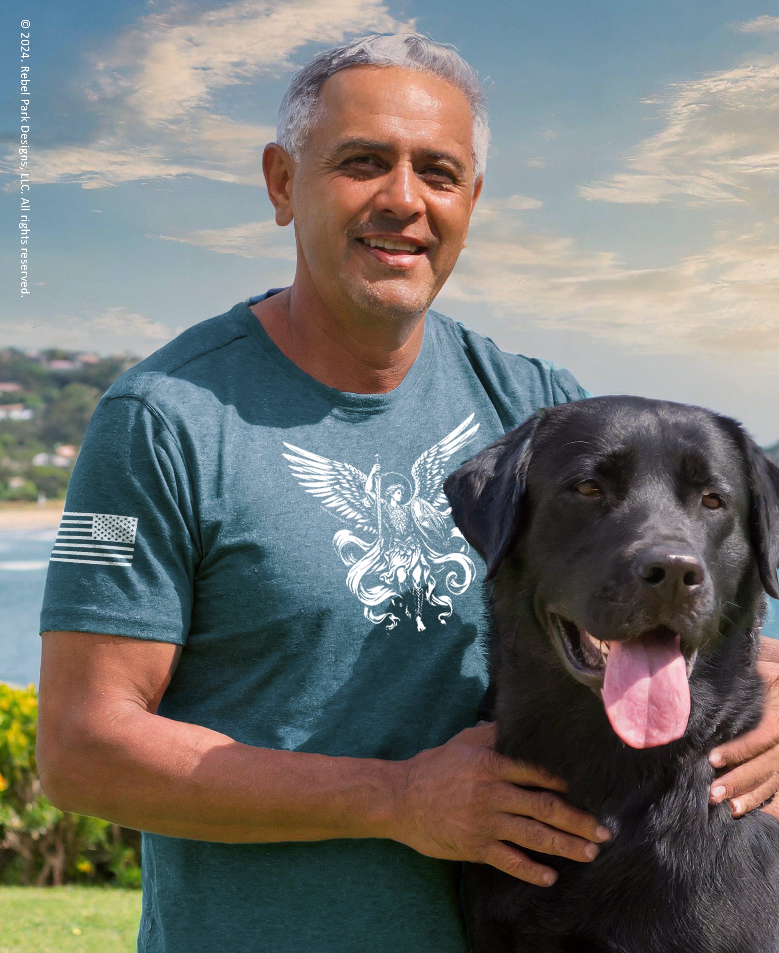 Man in a blue t-shirt with a dog on a grassy area with a beach and sky in the background