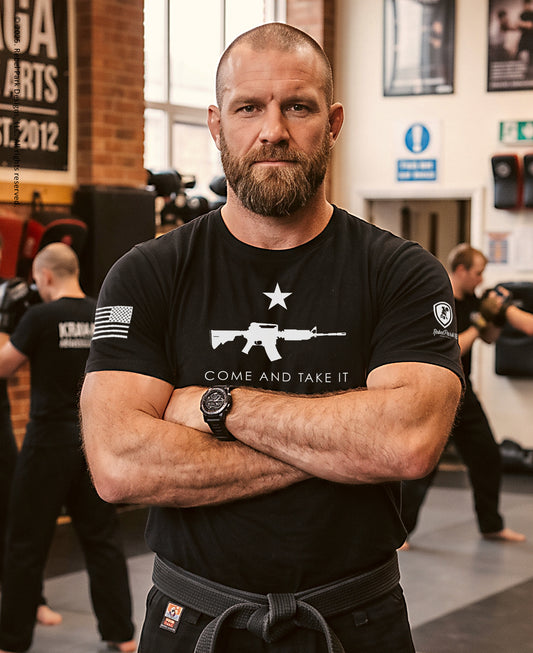 A muscular man with a beard wearing a black t-shirt with a white rifle graphic and the words 'COME AND TAKE IT' stands with his arms crossed in a martial arts dojo. The background shows other people training and gym equipment.