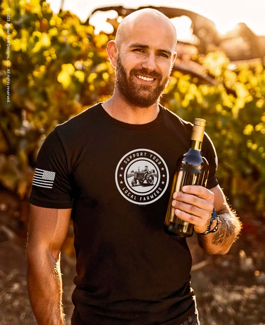 Smiling man in black Support Your Local Farmers T-shirt holding a bottle of wine in a vineyard.