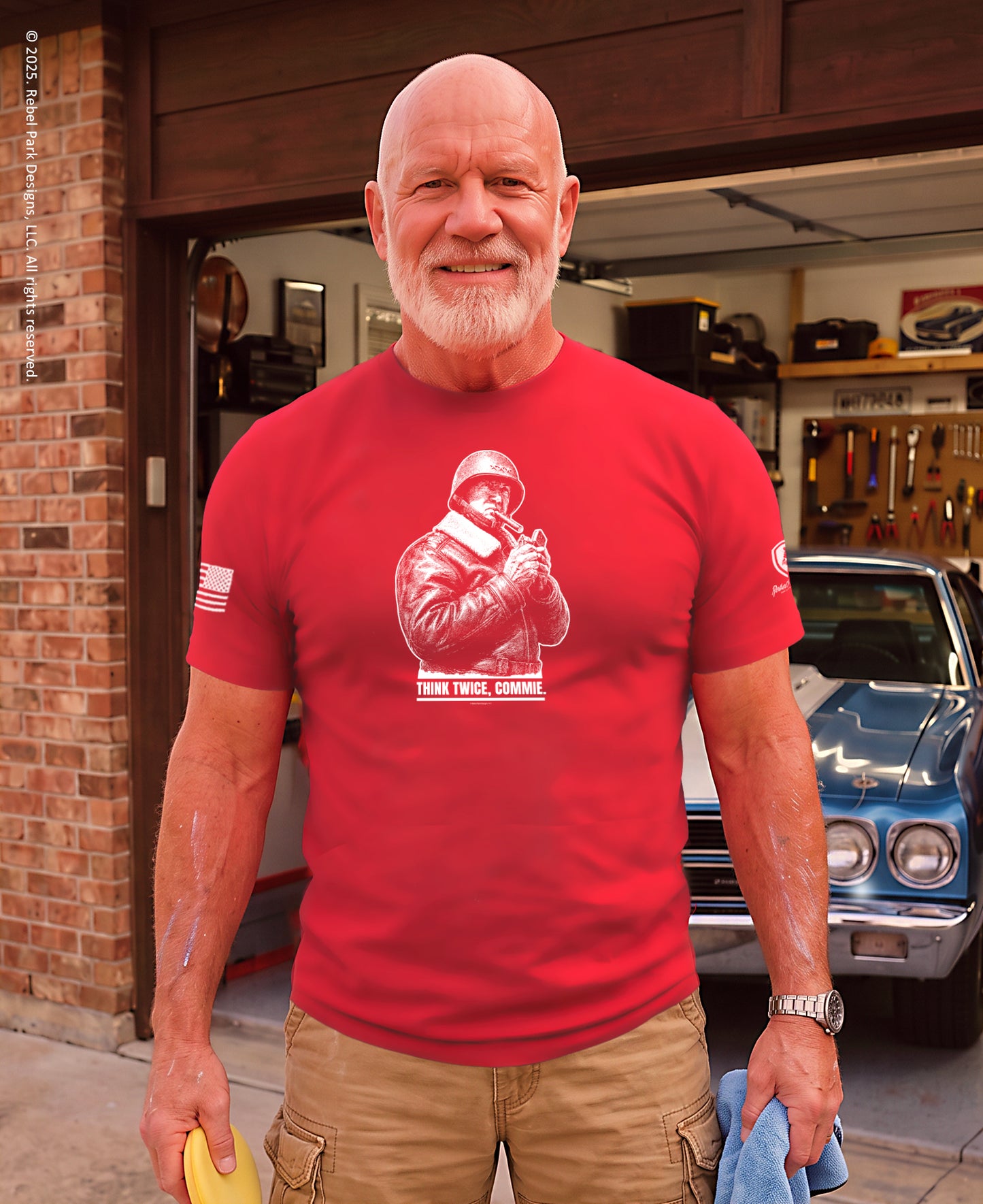 A smiling older man with a white beard stands in a garage wearing a bright red graphic T-shirt with a white military-style print and the words 'THAT TRUCK, GUNNISON' on the front. A blue classic car is visible behind him, along with a brick wall and garage interior.