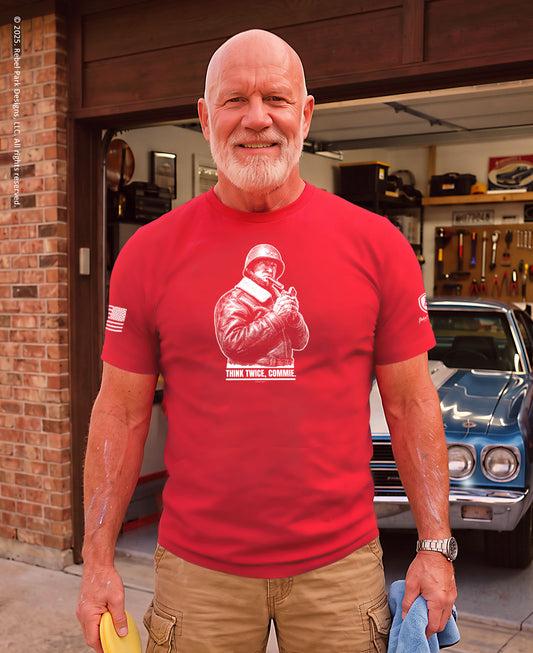 A man wearing a bright red graphic t-shirt with a white print of a soldier and the text 'THREE TIMES GUNNER' stands in front of an open garage door with a blue classic car in the background.