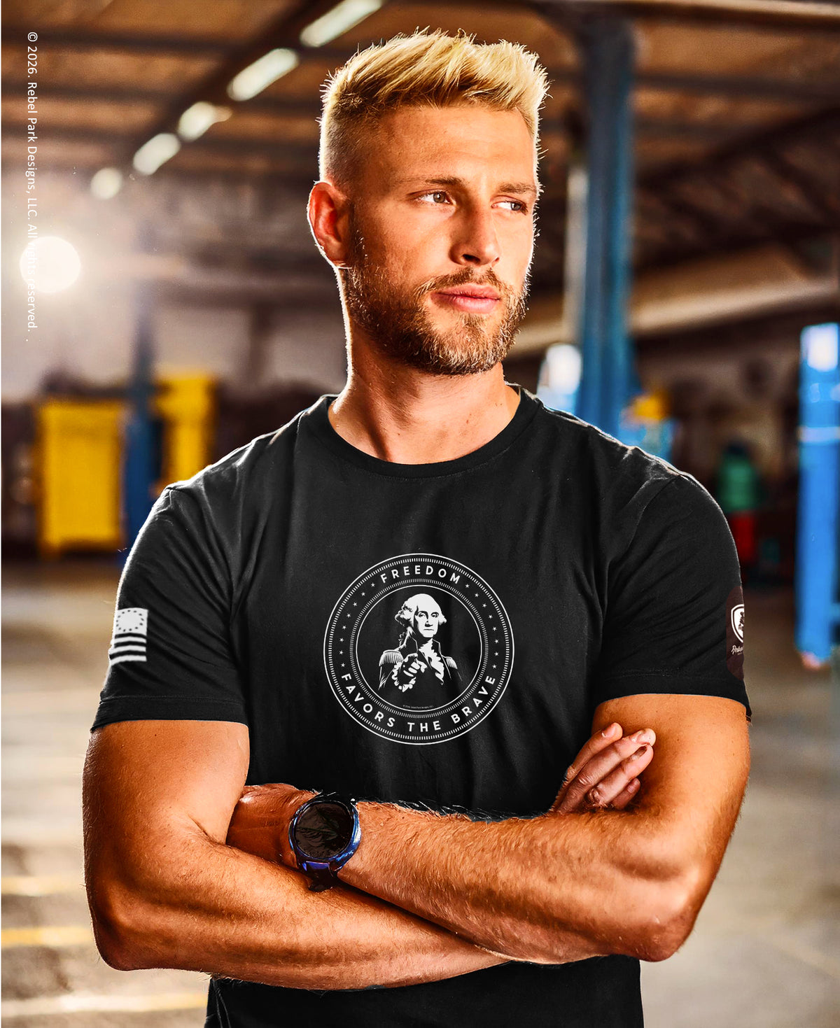 A muscular man with blond hair and a beard stands with arms crossed in an industrial garage setting, wearing a black t-shirt with a circular white graphic on the chest featuring a portrait and the text 'INDEPENDENCE BEFORE THE BRIDE'.
