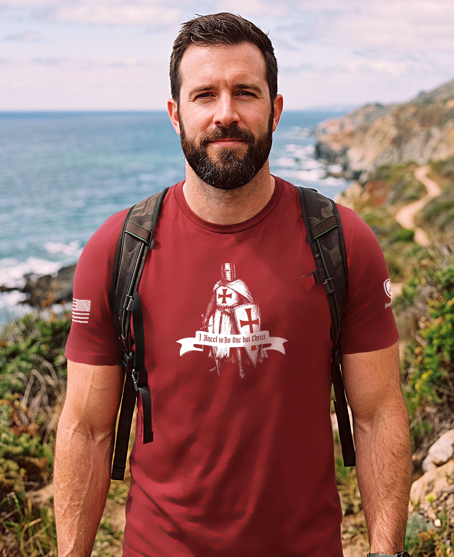 A man with a beard wearing a maroon t-shirt with a white cross and 'I PRAY IN THE DARK' graphic, standing outdoors on a rocky cliffside with the ocean in the background.