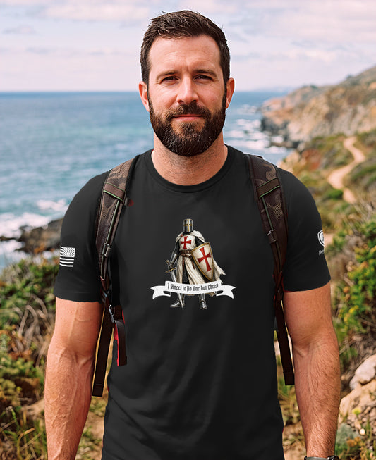 A man wearing a black graphic t-shirt with a Templar knight illustration and the text 'I swear to the Lord that I will obey', standing outdoors on a rocky cliffside path overlooking the ocean.