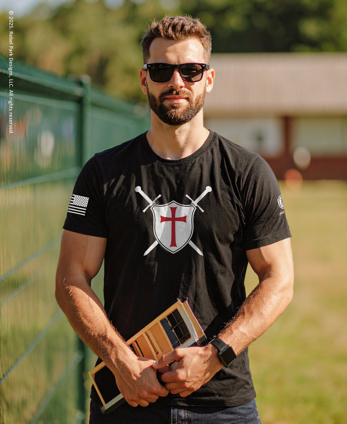Man wearing a black t-shirt with a logo, holding a wooden object outdoors.