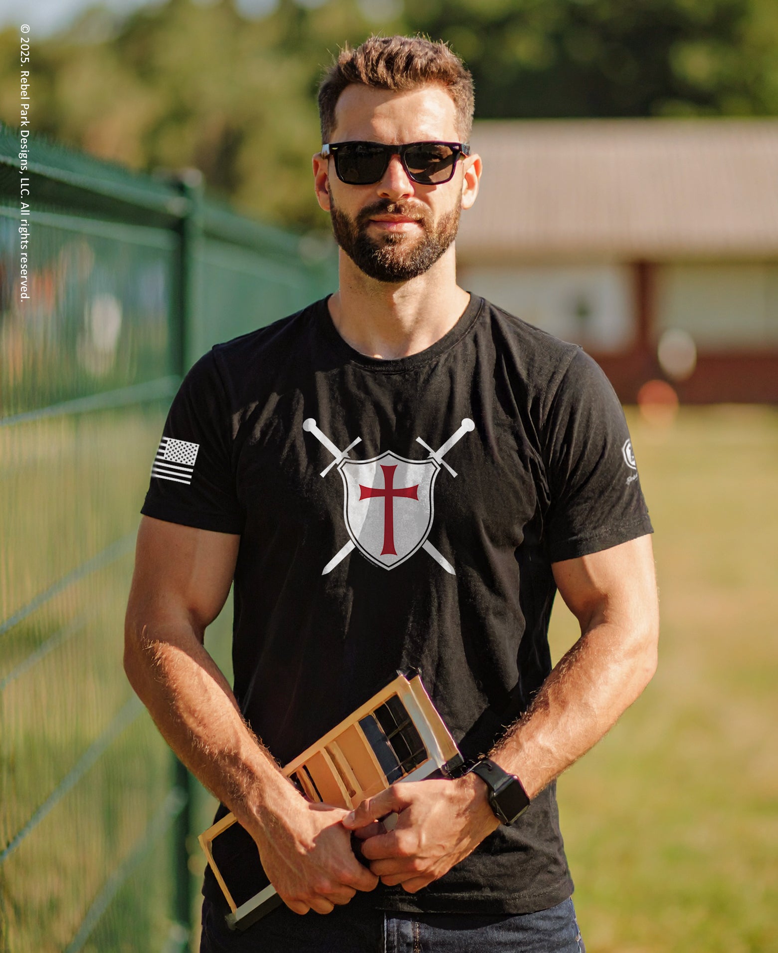 Man wearing a black t-shirt with a logo, holding a wooden object outdoors.