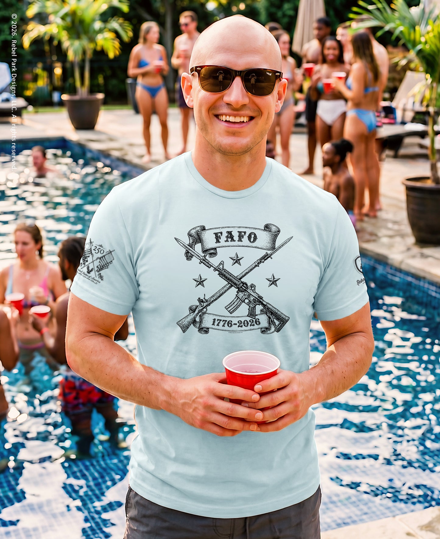 Man wearing a light blue t-shirt with a graphic design, holding a red cup, standing by a pool with people in the background.
