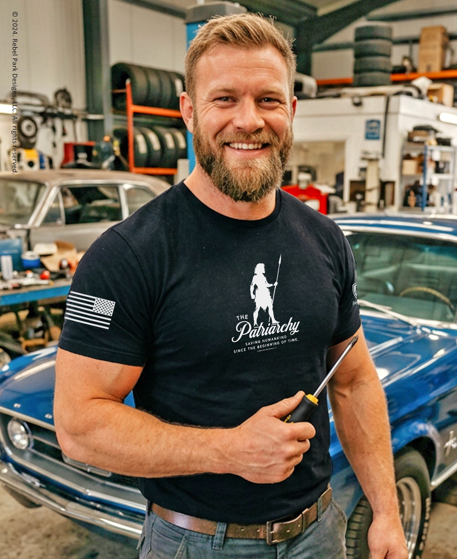 Man in a garage wearing a black t-shirt with a logo, holding a tool.
