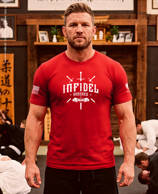 A muscular man wearing a red graphic t-shirt with 'INFIDEL ARMAGED' printed in white on the chest, standing in a martial arts dojo with other practitioners blurred in the background.