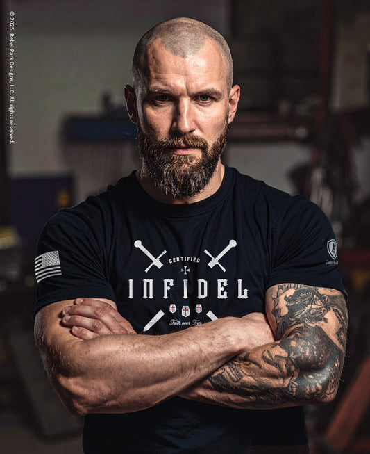 Man wearing a black t-shirt with 'Infidel' text and crossed swords design, standing in a workshop.