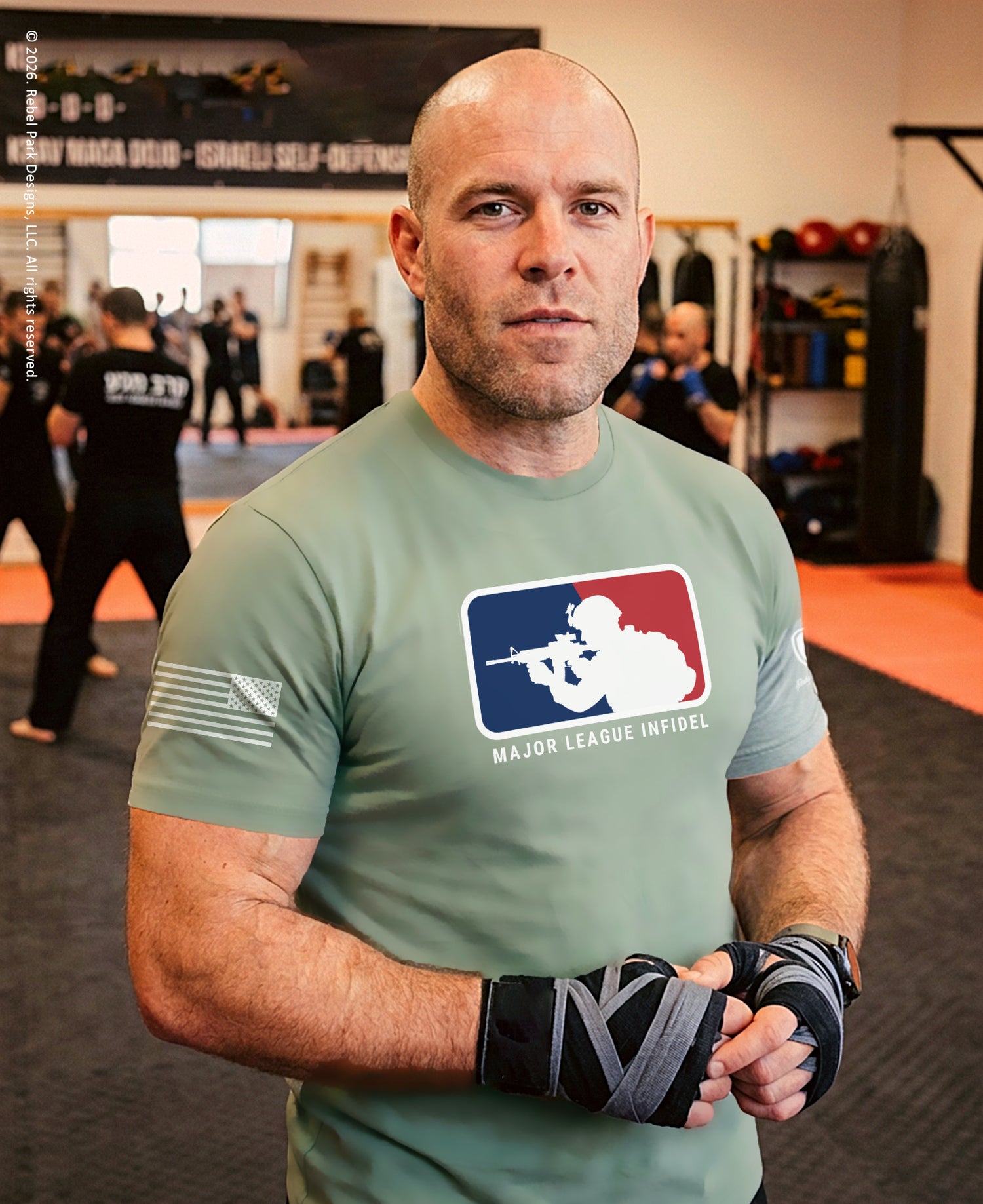 A bald man in a gym wearing a sage green t-shirt with a 'Major League Infuse' logo on the chest, black and gray hand wraps, and looking directly at the camera. The background shows a martial arts or training gym with other people and equipment.