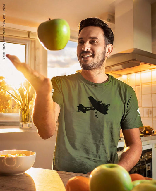 Man in olive green B-17 bomber graphic tee playfully tossing a green apple in a bright kitchen.