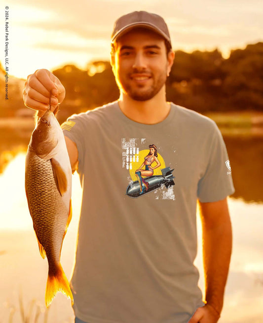 Man holding a caught fish while wearing a humorous t-shirt with fishing graphic art at sunset.