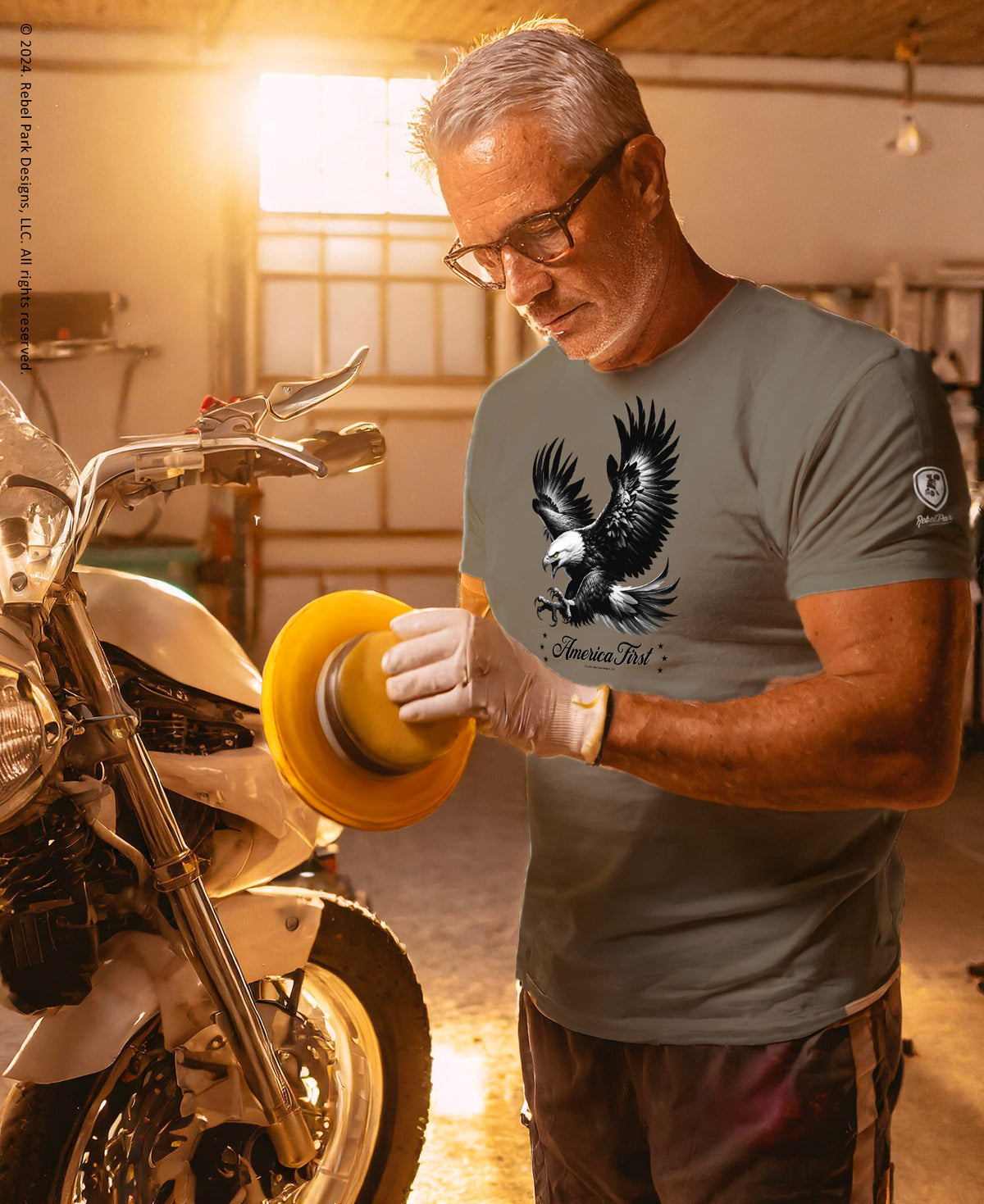 A man wearing a gray t-shirt with a black eagle graphic and the words 'American Iron' stands in a workshop next to a motorcycle, holding a yellow sanding disc.