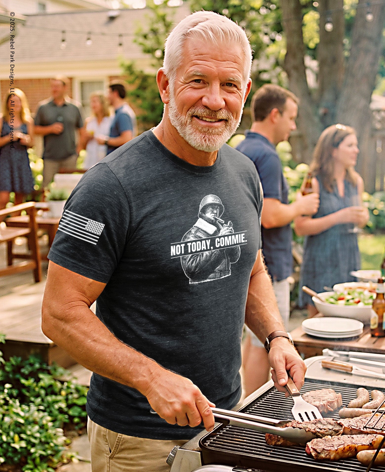 A smiling man with white hair and a beard grilling outdoors at a backyard barbecue while wearing a dark heather gray graphic t-shirt with a white graphic print and text.