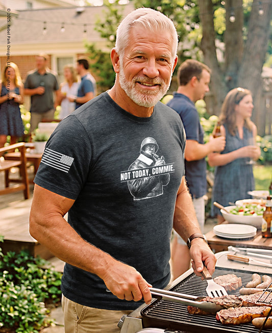 A smiling man with white hair and a beard grilling outdoors at a backyard barbecue while wearing a dark heather gray graphic t-shirt with a white graphic print and text.
