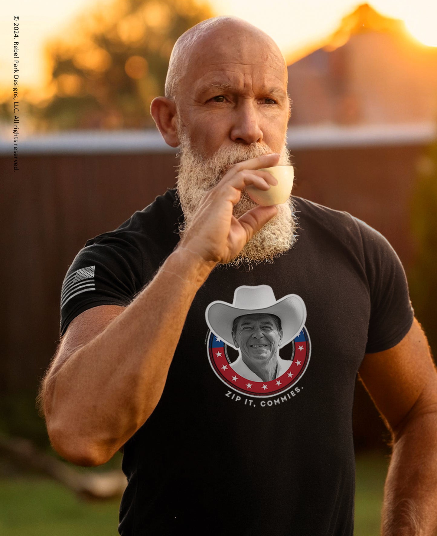 A muscular bald man with a white beard wearing a black graphic t-shirt, holding a white apple near his mouth in an outdoor setting during golden hour. The t-shirt features a circular graphic of a smiling man in a cowboy hat with the text 'ZIP IT.