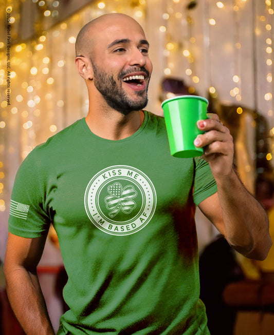 A smiling bald man with a beard holds a bright green plastic cup while wearing a green graphic t-shirt with a circular 'Kiss Me I'm Based Here' shamrock design. The background features warm, out-of-focus string lights.
