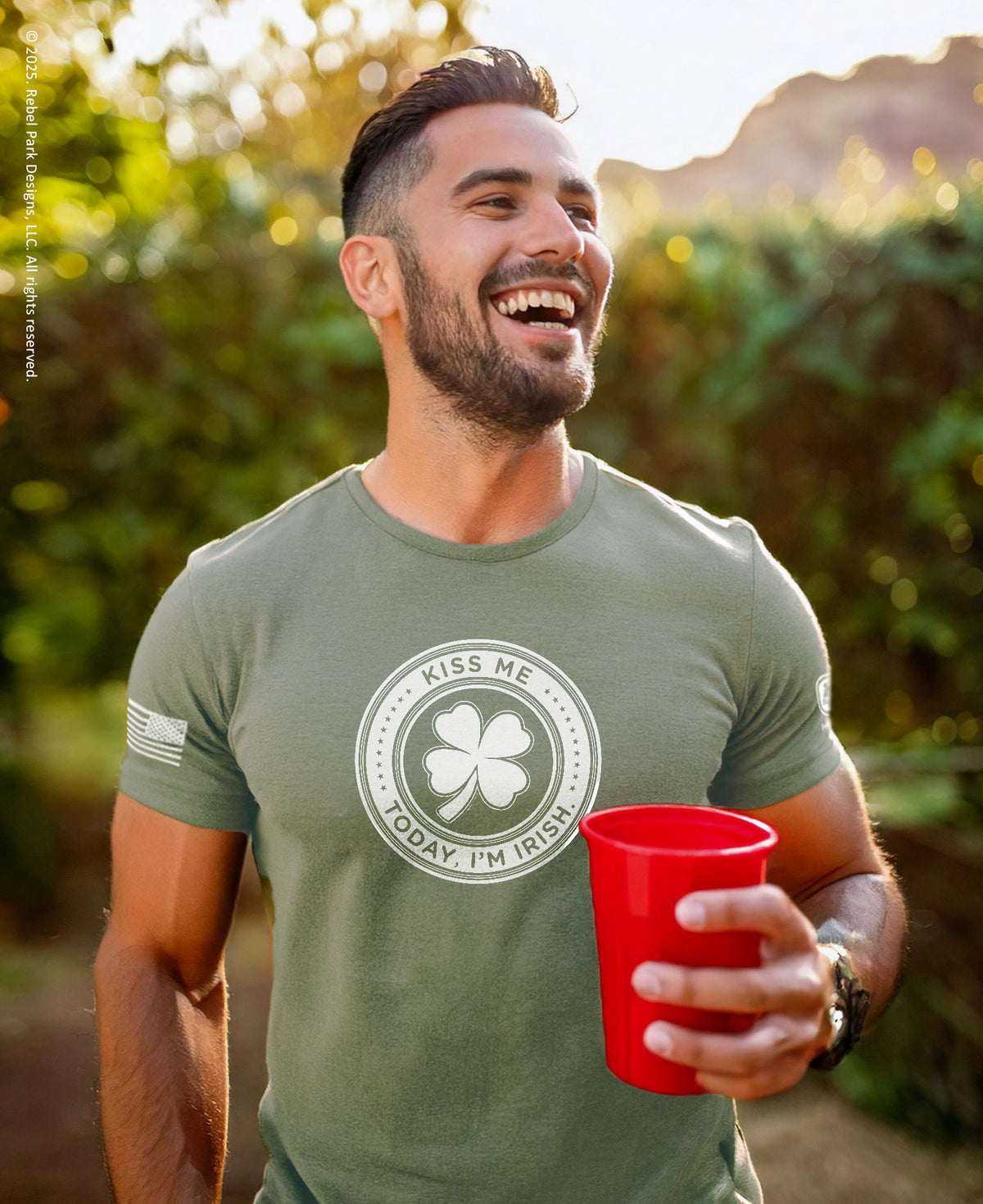 A smiling man outdoors wearing an olive green t-shirt with a circular graphic that reads 'KISS ME TODAY I'M IRISH' around a shamrock, holding a red plastic cup.