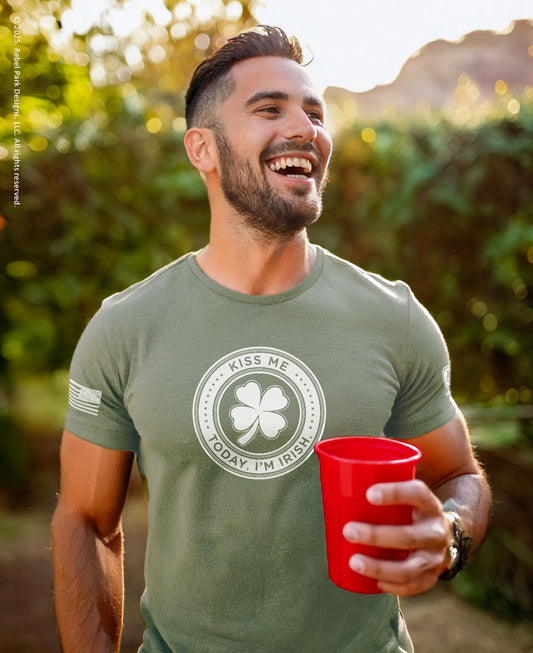 A smiling man outdoors wearing an olive green t-shirt with a circular graphic that reads 'KISS ME TODAY I'M IRISH' around a shamrock, holding a red plastic cup.