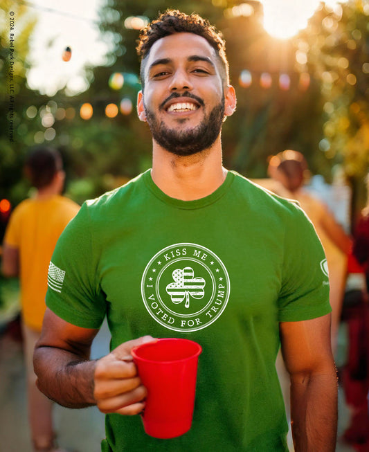 A smiling man outdoors at sunset wearing a green crew-neck t-shirt with a white circular graphic that reads 'KISS ME VOTED FOR THEIR' around a shamrock design. He is holding a red plastic cup.