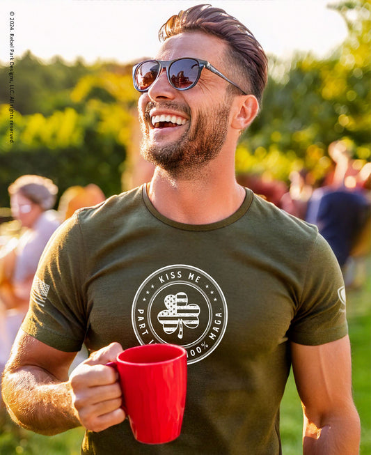 A man outdoors wearing an olive green graphic t-shirt with a white circular 'Kiss Me Paris Don't Bo' design, holding a red mug. He is wearing sunglasses and smiling, with a blurred green background.