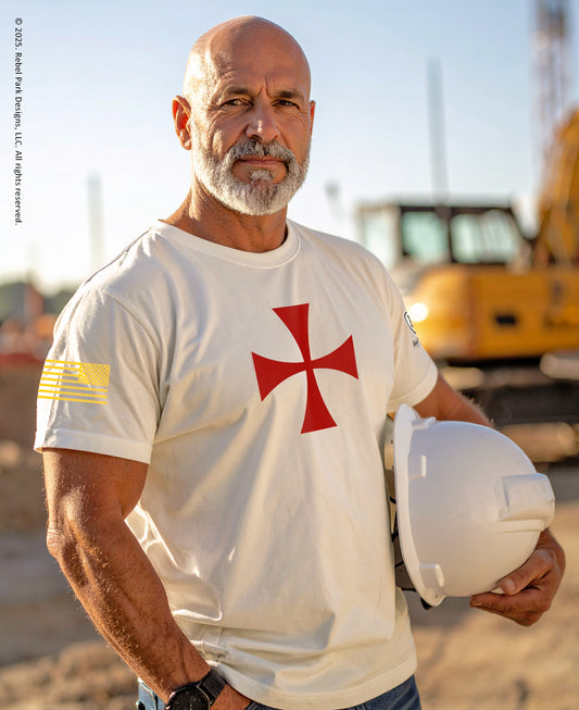Man wearing a white t-shirt with a red cross logo, holding a hard hat on a construction site.