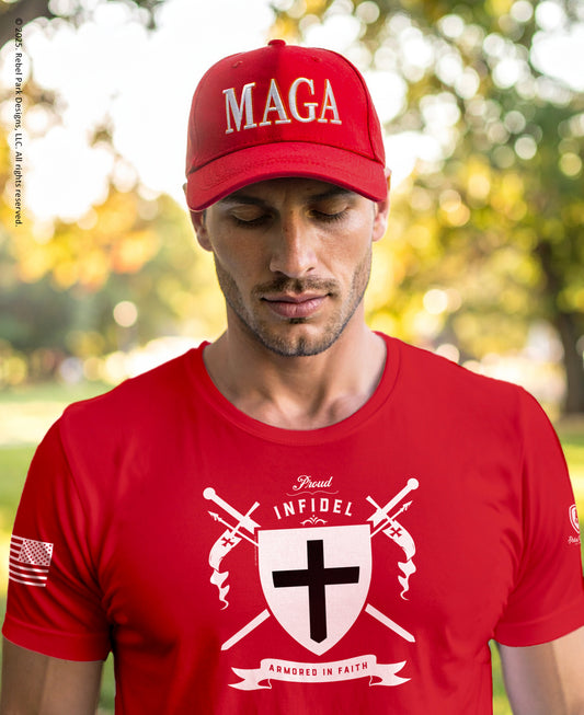 A man wearing a red baseball cap with 'MAGA' in white and a matching red t-shirt with a white religious graphic featuring a cross, swords, and the words 'BLOOD INFIDEL ARMED IN FAITH'. The background is a blurred outdoor park scene.
