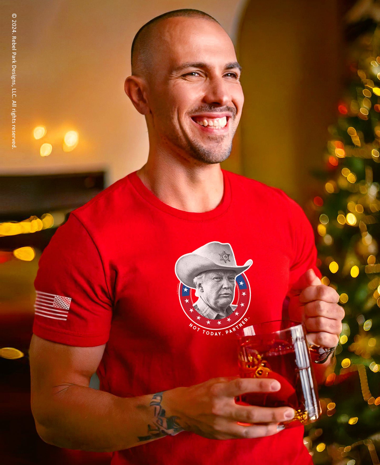 Man wearing a red t-shirt with a graphic design, holding a glass of wine, with a blurred Christmas tree in the background.