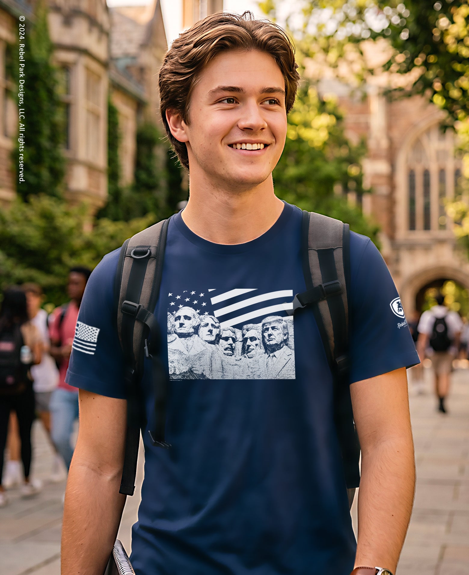 A smiling young man wearing a navy blue graphic t-shirt with a monochrome American flag and portrait print, standing outdoors on a campus walkway with a backpack on.