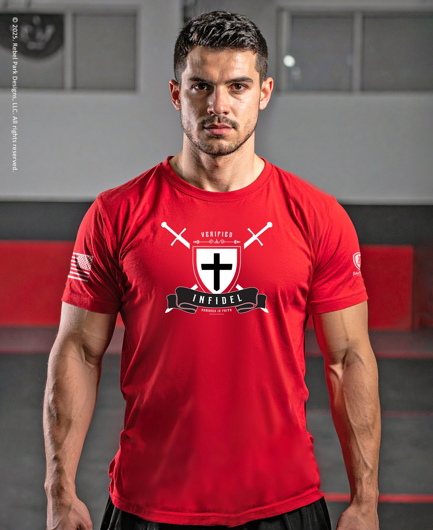 A man wearing a bright red athletic t-shirt with a black and white cross graphic and the text 'VERIFIED INFIDEL' on the chest, set against a gym background.